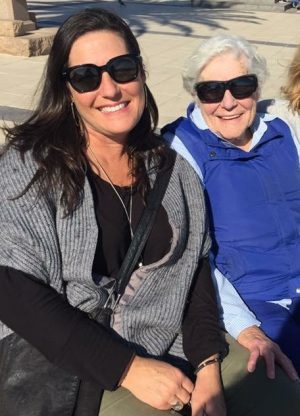 An elderly mother in a blue jacket with white hair and dark sunglasses sit smiling next to her grown daughter who is also smiling wearing dark sunglasses and a grey jumper. They are happy that the mother can stay in her own home and be safe thanks to the Ageing In Place system they have.