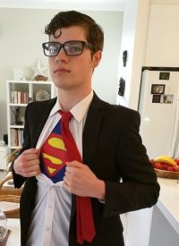 A young man with dark hair and coke-bottle glasses opens his shirt to reveal the S of the superman costume that he wears underneath, he is a safety superhero to those who wish to live at home in their later years with the Ageing In Place system.